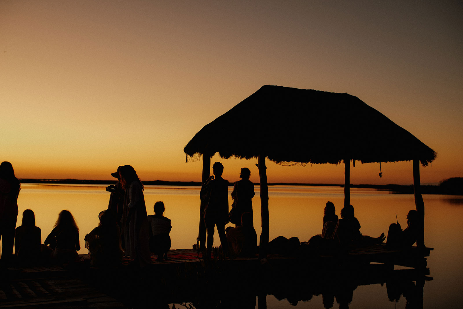 A group gathered under a palapa at sunset by the water